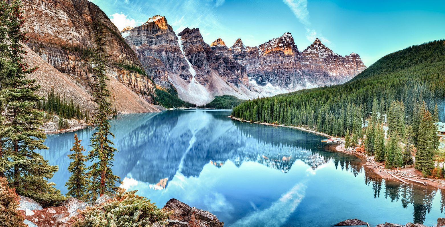 Vue panoramique du lac Moraine, situé dans le parc national de Banff en Alberta, entouré de montagnes majestueuses, de forêts denses et de l'eau turquoise cristalline sous un ciel bleu clair.